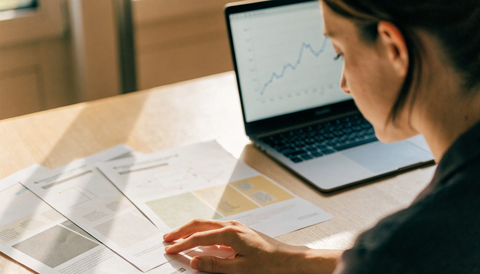 A woman reviewing printed presentation slides spread across a desk beside an open laptop showing a line chart.